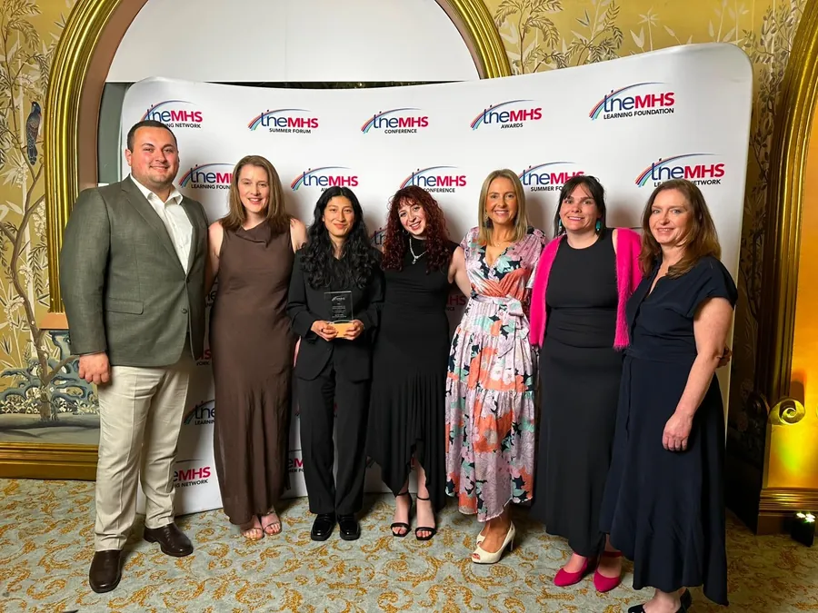 The Youth Peer Work Team, seven people, holding their glass TheMHS Award trophy in front of the TheMHS branded media wall