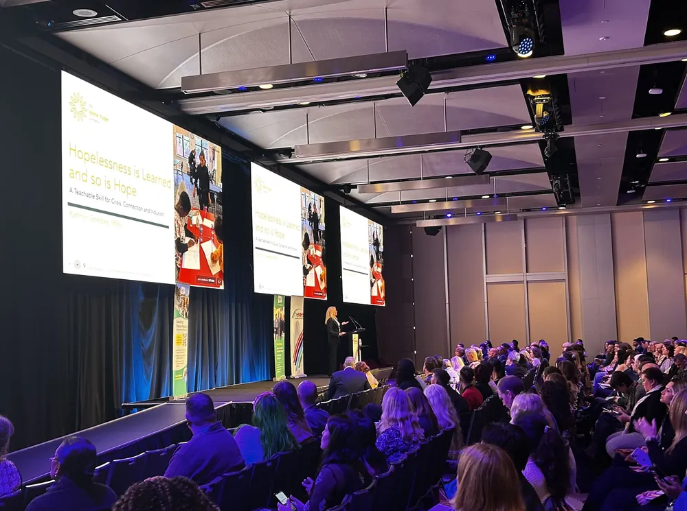 Kathryn Goetzke delivering the opening keynote to a packed room, with three large screens reading Hopelessness is Learned and so is Hope