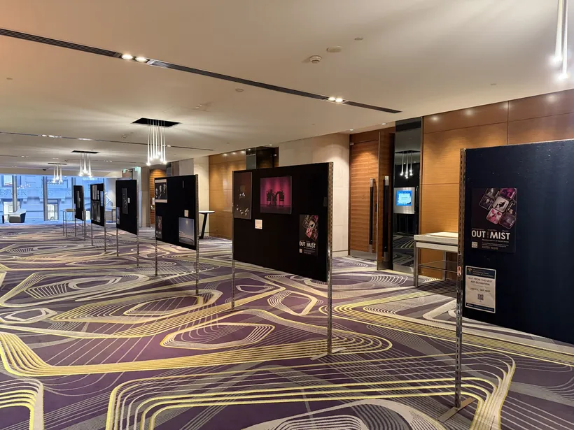 Out From The Mist exhibition in a hotel foyer: framed photographs on black display boards along a patterned carpet