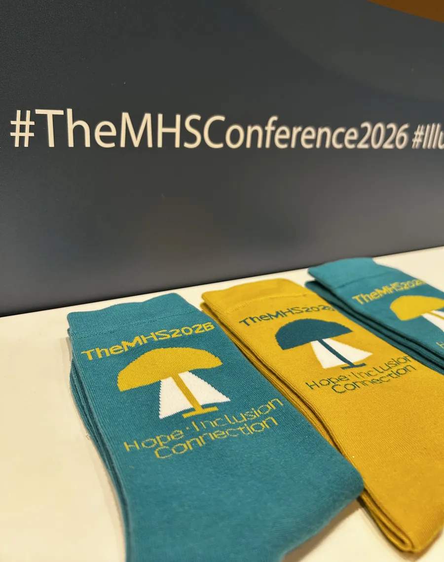 Teal and gold TheMHS 2026 conference socks laid out in front of a wall reading hashtag TheMHSConference2026